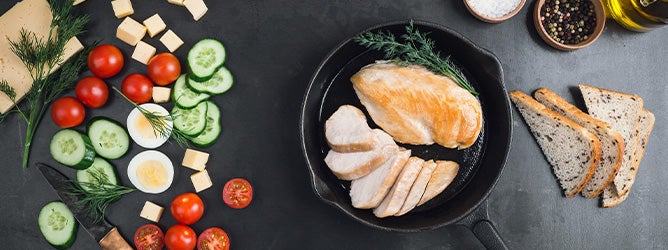 Sartén con pollo sobre una mesa que tiene vegetales, trozos de queso, sal, pimienta y huevo